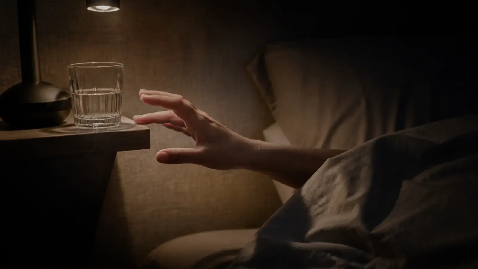 hand reaching sideways in a dark room toward a narrow bedside surface