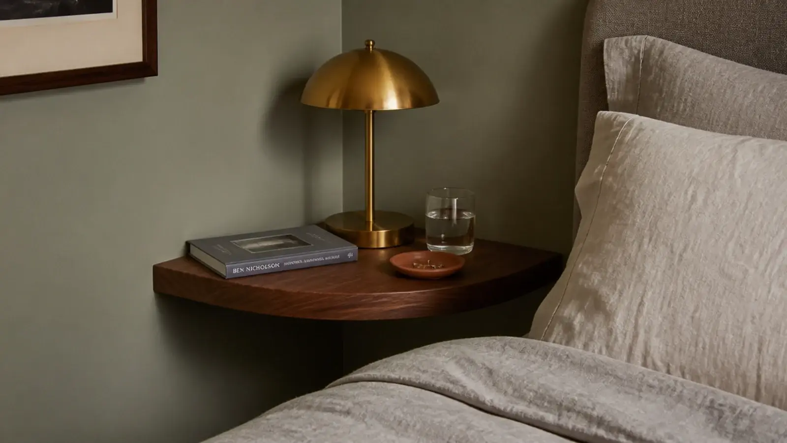 small floating corner shelf beside a bed holding a book lamp and water glass