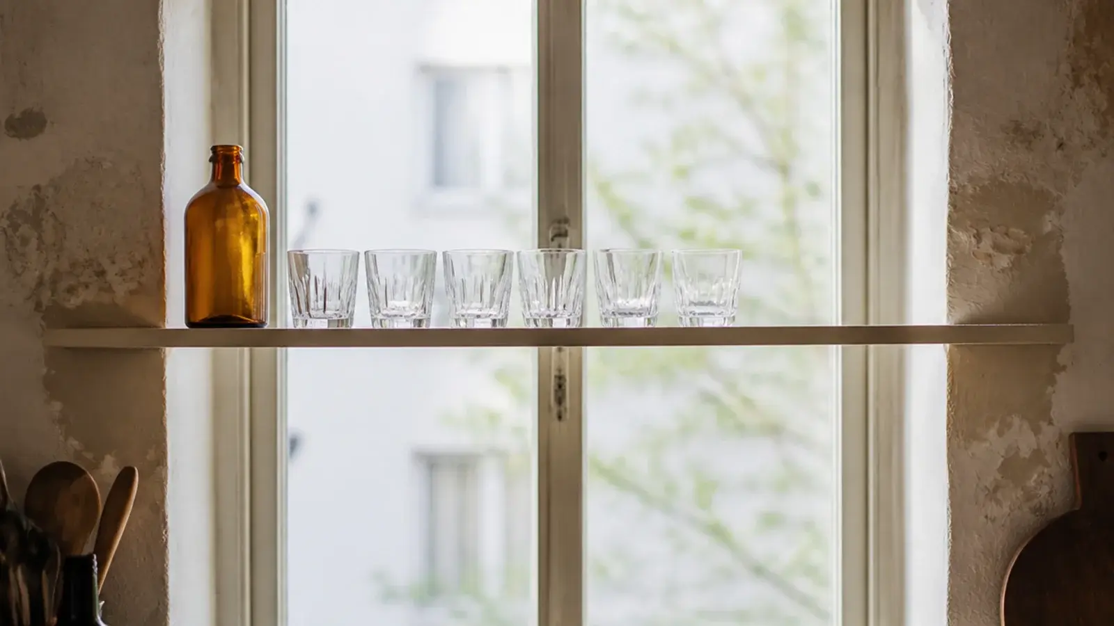 slim kitchen shelf crossing a tall window holding backlit glassware