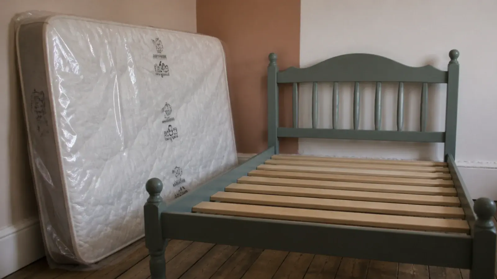 new wrapped single mattress leaning beside a secondhand painted bed frame