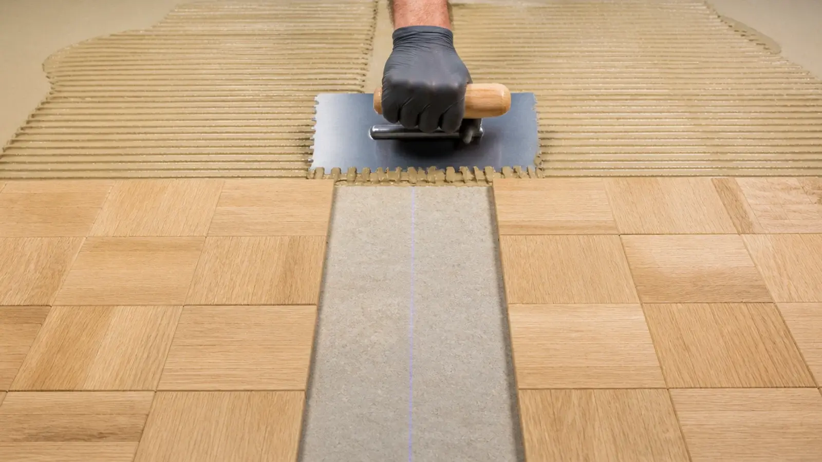 notched trowel spreading adhesive beside dry laid oak parquet blocks